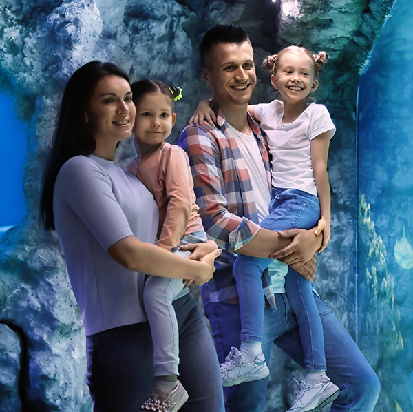 A family of four posing together in front of a glowing blue aquarium tunnel