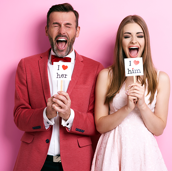 A couple in formal dress laughing behind I-love-her and I-love-him photo booth props on a pink backdrop