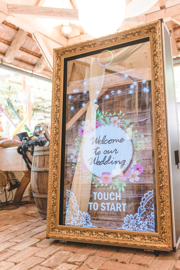 A mirror photo booth in an ornate gold frame standing in a timbered barn, its screen showing a floral Welcome to our Wedding start screen beside a barrel of photo props.