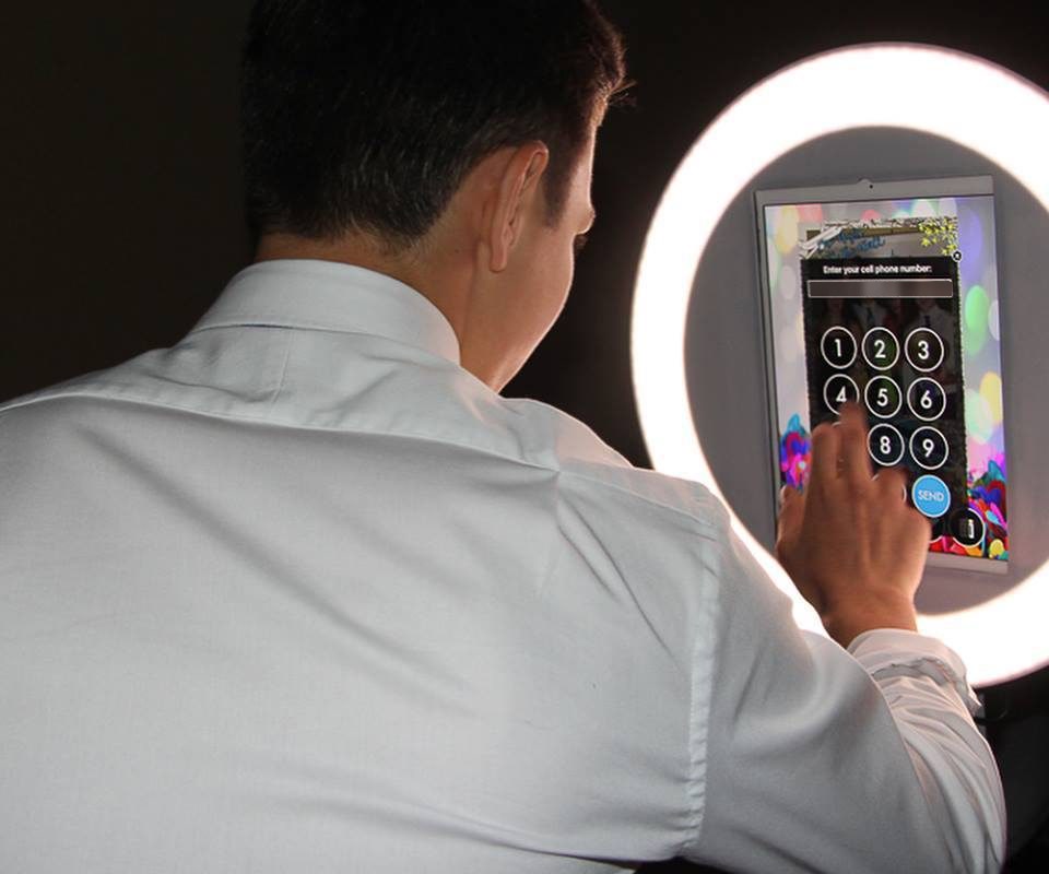 A guest at an evening event taps his mobile number into the glowing screen of a ring-lit Curator iPad photo booth, with the numeric keypad and blue SEND button ready to text him the photo he has just taken.